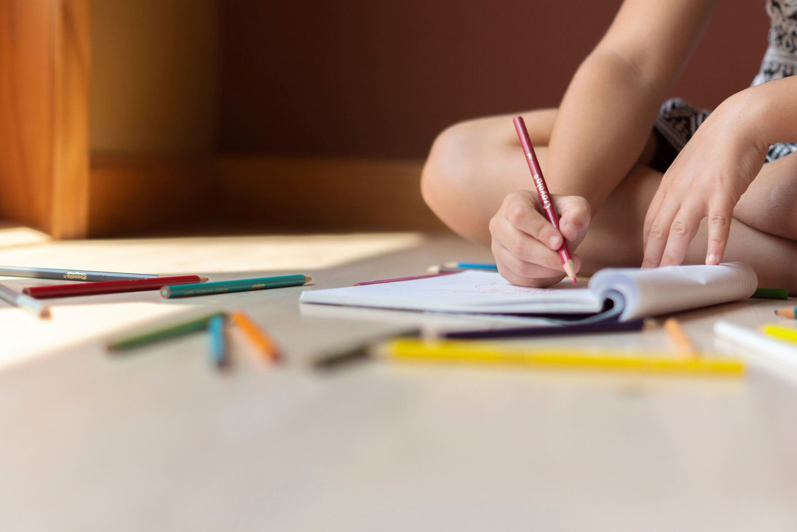 Child drawing in a notebook with colorful pencils on the floor